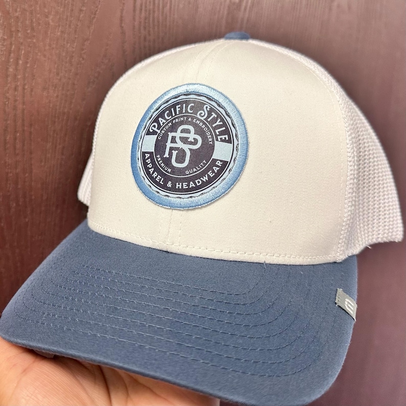 May include: A white and blue baseball cap with a mesh back. The front of the cap features a blue and white circular patch with the text "Pacific Style Custom Print & Embroidery Premium Quality Apparel & Headwear".