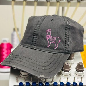 May include: A distressed charcoal gray baseball cap featuring a pink embroidered llama design. The cap has a curved brim, a top button, and visible stitching details. The hat appears to be made of a soft, worn fabric.
