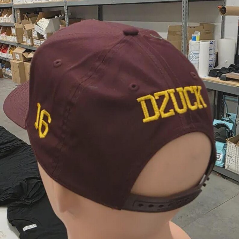 May include: A maroon baseball cap featuring the yellow embroidered text DZUCK on the back panel and the number 16 in yellow embroidery on the side panel. The cap includes a matching maroon adjustable plastic snapback closure at the rear.