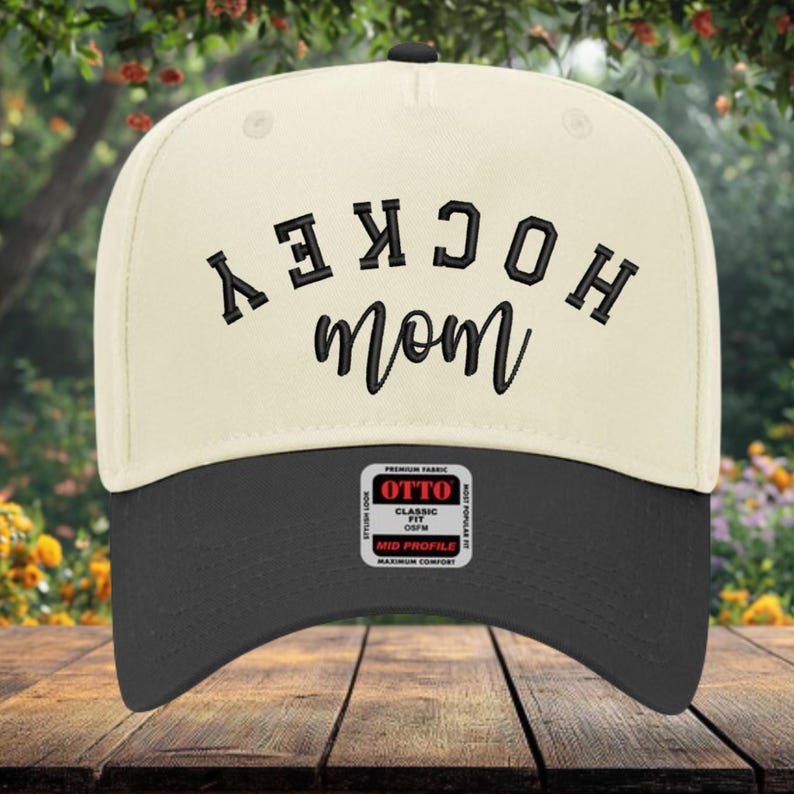 May include: A cream-colored baseball cap with a black brim featuring the embroidered text HOCKEY in a bold, arched block font and the word mom in a cursive script below it. A rectangular sticker with the text OTTO CLASSIC FIT OSFM is visible.