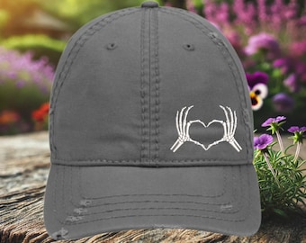 May include: A gray distressed baseball cap with a white skeleton hand design making a heart shape.
