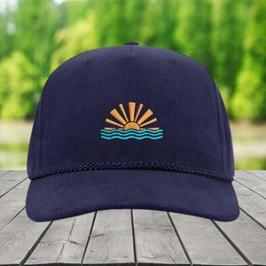 May include: A navy blue baseball cap featuring an embroidered design of a sun rising over turquoise waves. The cap has a curved peak and a braided rope detail. The background shows blurred green trees and water.