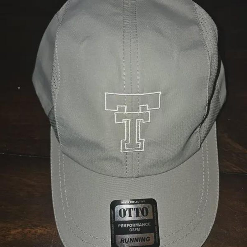 May include: A light gray baseball cap featuring a white embroidered double T logo on the front panel. The cap includes side mesh ventilation panels and a black sticker on the brim with the text OTTO PERFORMANCE OSFM RUNNING.