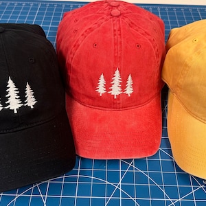 May include: Three baseball caps in black, red, and yellow with a white embroidered design of three pine trees on each.