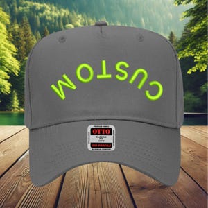 May include: A grey baseball cap featuring the word CUSTOM embroidered in bright lime green thread in an arched orientation across the front panels. A rectangular black and white product label is attached to the centre of the cap.