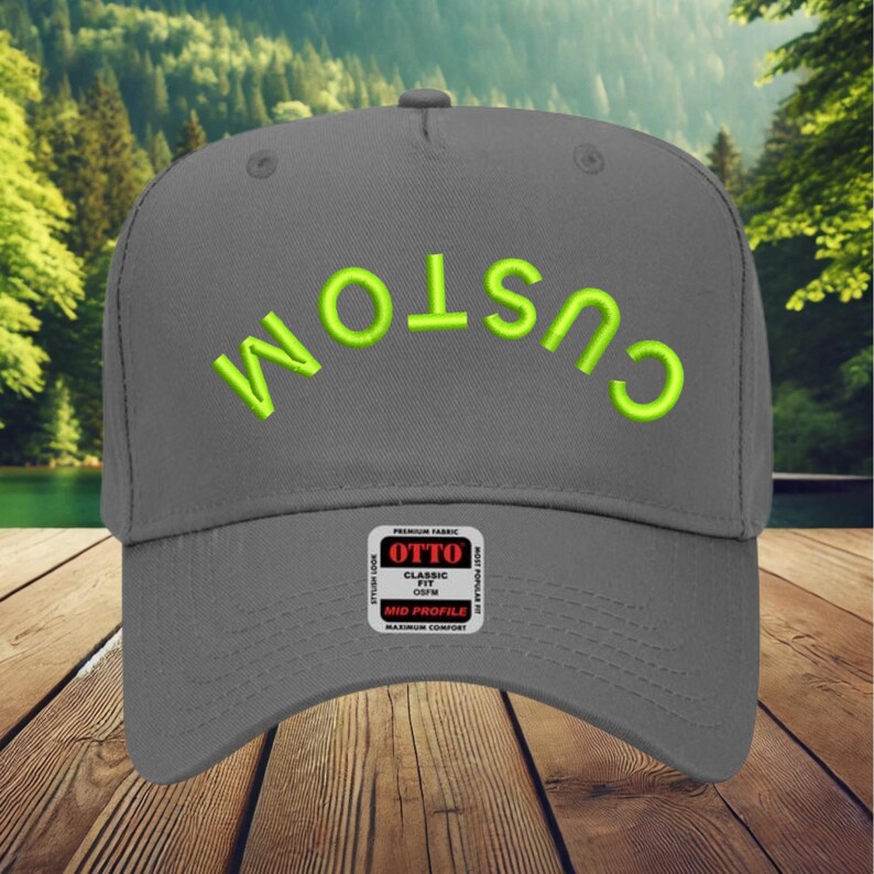 May include: A grey baseball cap featuring the word CUSTOM embroidered in bright lime green thread in an arched orientation across the front panels. A rectangular black and white product label is attached to the centre of the cap.