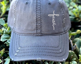 May include: A distressed gray baseball cap with the word "faith" embroidered in white script on the side. The cap has a curved brim and a worn, vintage look, with visible stitching and distressed edges.