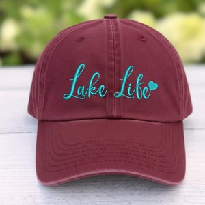 以下が含まれることがあります： マルーン色の野球帽。ターコイズ色の筆記体で「Lake Life」の文字が刺繍されています。「Life」の横にはターコイズ色のハートがあります。帽子にはカーブしたつばと布製のストラップが付いています。