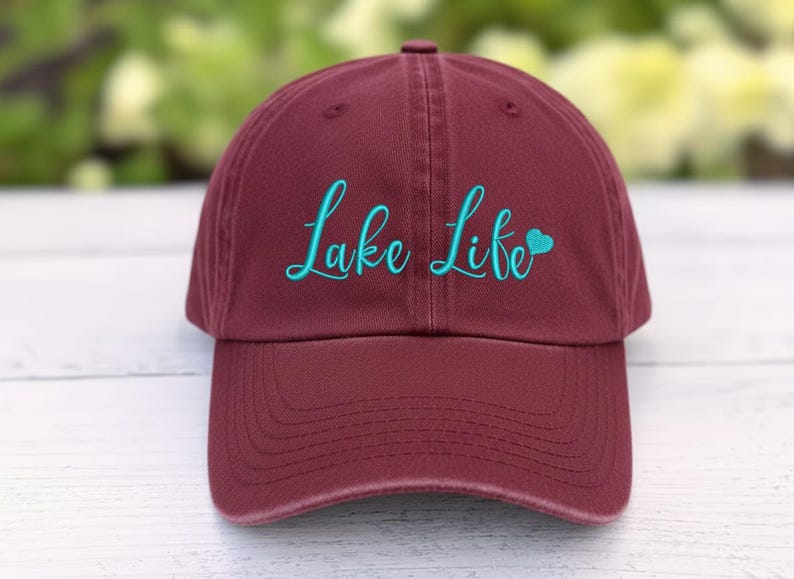 以下が含まれることがあります： マルーン色の野球帽。ターコイズ色の筆記体で「Lake Life」の文字が刺繍されています。「Life」の横にはターコイズ色のハートがあります。帽子にはカーブしたつばと布製のストラップが付いています。