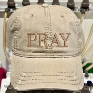 May include: A beige baseball cap with the word "PRAY" embroidered in a brown thread across the front. The cap has a distressed look with visible stitching and small holes. The hat is a classic six-panel design.