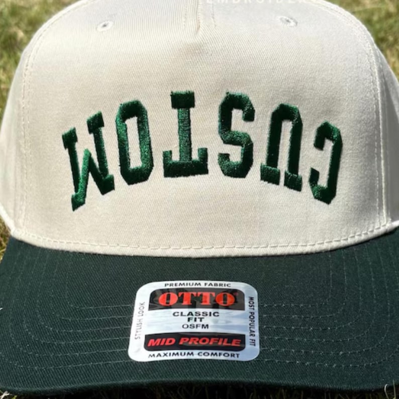 May include: A cream-coloured baseball cap with a dark green brim and dark green embroidered text that reads CUSTOM in an arched font. A rectangular sticker on the brim features the text OTTO, PREMIUM FABRIC, CLASSIC FIT, OSFM, and MID PROFILE.