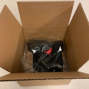 May include: A black baseball cap with a red cardinal bird logo inside a cardboard box.