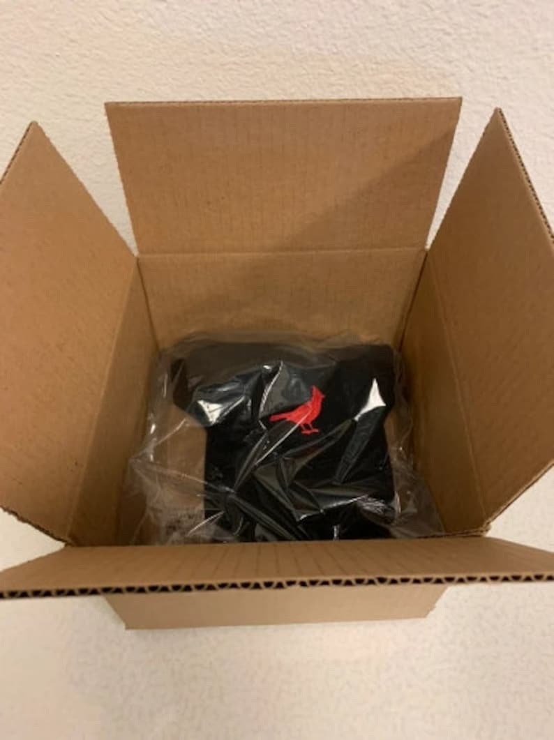 May include: A black baseball cap with a red cardinal bird logo inside a cardboard box.