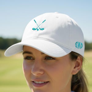 Peut inclure: Casquette de baseball blanche avec une visière incurvée, ornée d'un motif brodé turquoise de clubs de golf croisés et d'une balle. La casquette porte également des initiales turquoise brodées sur le côté. En arrière-plan, un parcours de golf vert flou.