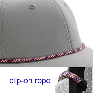 May include: A light grey baseball cap featuring a decorative twisted rope trim in red, white, and blue colours. The cap includes a detachable clip-on rope accessory, as indicated by the text clip-on rope, which attaches to the side of the hat.