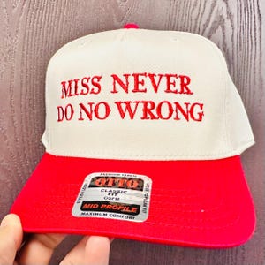 May include: A white and red baseball cap with the embroidered text "MISS NEVER DO NO WRONG". The cap has a red brim and a white crown. The cap has a "OTTO" label.