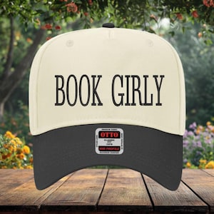 May include: A white and black baseball cap with the text "BOOK GIRLY" embroidered on the front. The cap has a black brim and a white crown. The cap has a label on the back that says "OTTO CLASSIC FIT MID PROFILE MAXIMUM COMFORT".