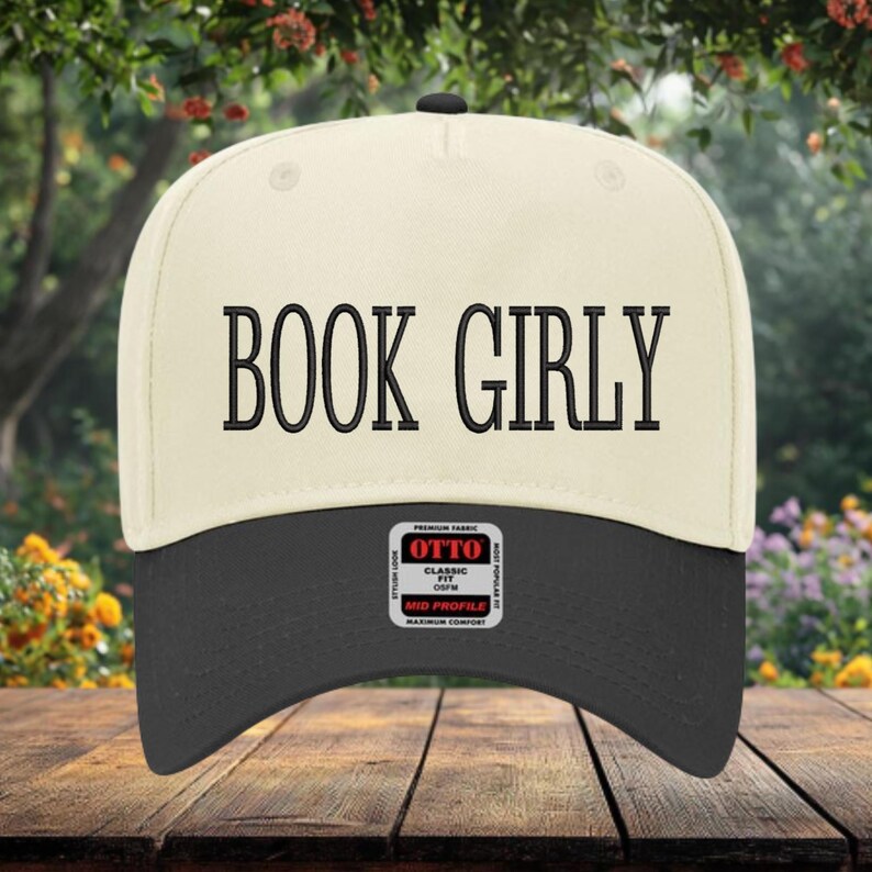May include: A white and black baseball cap with the text "BOOK GIRLY" embroidered on the front. The cap has a black brim and a white crown. The cap has a label on the back that says "OTTO CLASSIC FIT MID PROFILE MAXIMUM COMFORT".
