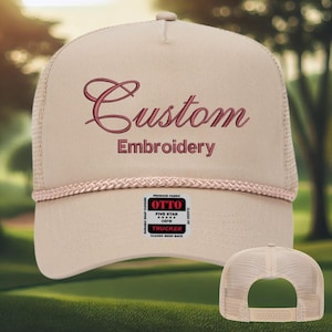 May include: A beige trucker hat featuring burgundy embroidered text reading "Custom Embroidery". The hat has a mesh back and a rope detail around the brim. A small label displays "OTTO" and "Trucker".