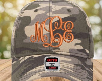 May include: Camouflage baseball cap with an embroidered orange monogram. The cap has a curved peak, a button on top, and a tag that reads "OTTO Classic Fit Camo". The hat is made of a premium fabric for maximum comfort.