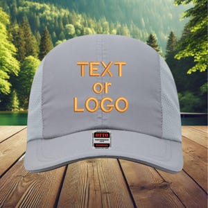 May include: A gray running cap with a curved brim and mesh side panels. The cap features embroidered orange text that reads "TEXT or LOGO". A small tag with the brand name "OTTO" is visible on the front.