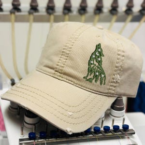 May include: A tan baseball cap with a distressed brim. The cap features an embroidered design of a wolf howling at the moon and stars in green thread. The cap is sitting on a white machine.