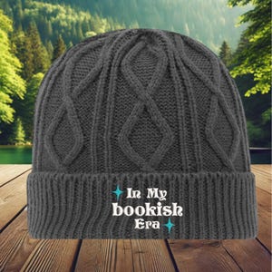 May include: Gray cable knit beanie with a white embroidered design that says "In My Bookish Era" with two small blue stars on either side of the text.
