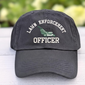 May include: A charcoal gray baseball cap featuring white embroidered text that reads LAWN ENFORCEMENT OFFICER in an arched design. Centered between the words is a small, detailed green and white embroidered graphic of a push lawn mower.