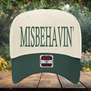 May include: A cream and forest green baseball cap with the word "MISBEHAVIN'" embroidered in green. The cap has a curved brim and a tag that reads "OTTO CLASSIC FIT OSFM MID PROFILE MAXIMUM COMFORT".