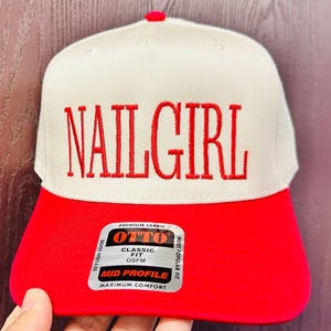 May include: A beige and red baseball cap with the text "NAILGIRL" embroidered in red on the front. The cap has a classic fit and a mid profile. The label on the side reads "OTTO" and "MOST POPULAR FIT".