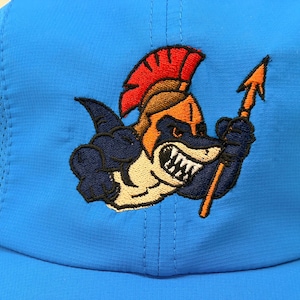 May include: A bright blue baseball cap featuring an embroidered graphic of a shark wearing a golden gladiator helmet with a red crest. The shark is depicted with sharp teeth and is holding a golden spear in its fin. The cap has mesh side panels.