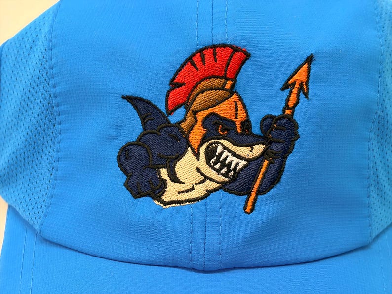 May include: A bright blue baseball cap featuring an embroidered graphic of a shark wearing a golden gladiator helmet with a red crest. The shark is depicted with sharp teeth and is holding a golden spear in its fin. The cap has mesh side panels.