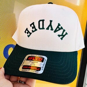 May include: A two-tone baseball cap featuring an off-white front panel and a dark green brim. The word KAYDEE is embroidered in dark green letters in an arched design on the front. A rectangular sticker on the brim reads OTTO CLASSIC FIT OSFM.