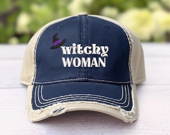 May include: A navy blue and beige baseball cap with a distressed, frayed brim. The front panel features an embroidered black witch hat with a purple band and the white embroidered text "witchy WOMAN" centered below the hat graphic.