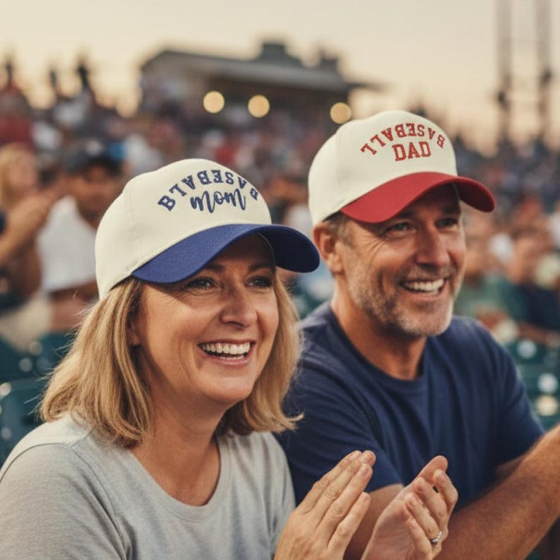 May include: Two baseball caps with off-white crowns. One cap features a blue brim and the text BASEBALL mom in blue. The other cap features a red brim and the text BASEBALL DAD in red. Both caps have a structured front panel and a curved brim.