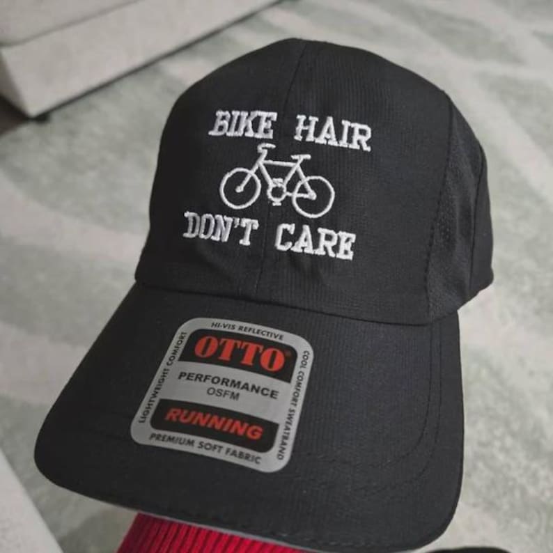 May include: A black baseball cap featuring white embroidered text that reads BIKE HAIR DON'T CARE with a bicycle graphic. The cap includes a silver and black sticker on the brim with the text OTTO PERFORMANCE OSFM RUNNING HI-VIS REFLECTIVE.