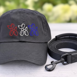 May include: A charcoal gray baseball cap embroidered with red, white, and blue balloon dog designs. A matching black dog leash with a padded handle and a metal clasp is next to the cap. The items are pet accessories.