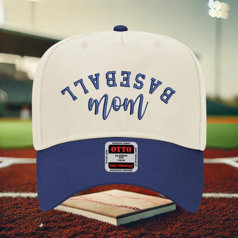 May include: A cream-colored baseball cap with a dark blue brim and dark blue embroidered text that reads BASEBALL mom. The cap features a central button on top and a rectangular sticker on the brim with the text OTTO CLASSIC FIT OSFM.