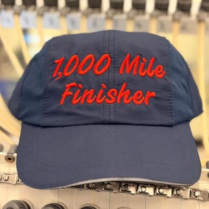 May include: A navy blue baseball cap featuring the embroidered red text 1,000 Mile Finisher on the front panel. The cap is constructed with a solid front panel and mesh side panels, showcasing a structured design with a curved brim and stitching.