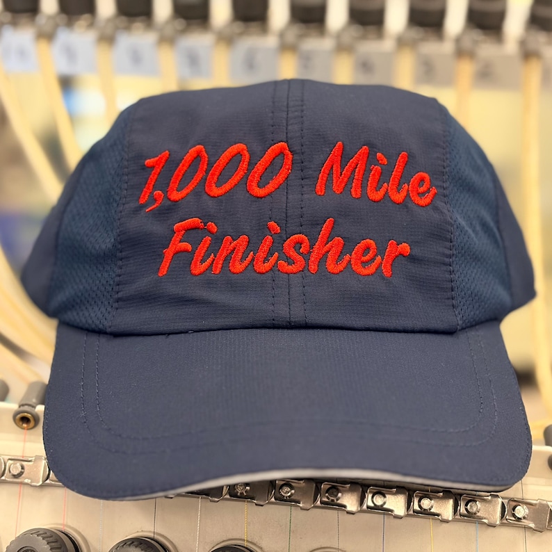 May include: A navy blue baseball cap featuring the embroidered red text 1,000 Mile Finisher on the front panel. The cap is constructed with a solid front panel and mesh side panels, showcasing a structured design with a curved brim and stitching.