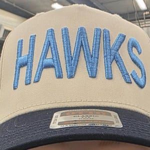 May include: A tan baseball cap with the word "HAWKS" embroidered in light blue across the front. The brim is navy blue. The cap has a classic fit and a tag that reads "MAXIMUM COMFORT".