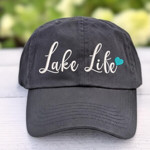 以下が含まれることがあります： 白い筆記体で「Lake Life」と刺繍された、小さなターコイズのハートが付いた濃い灰色の野球帽。 つばがカーブしており、柔らかい生地で作られています。