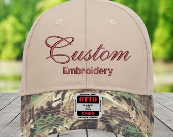 May include: A beige baseball cap with a camouflage pattern on the peak. The cap is embroidered with the word "Custom" above "Embroidery". The cap has an "OTTO" tag.