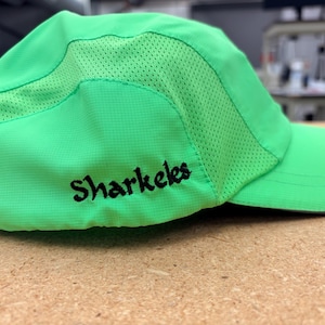 May include: A bright neon green baseball cap featuring a curved brim and breathable mesh side panels. The word Sharkeles is embroidered in black thread on the side of the cap. The hat is constructed from a lightweight, synthetic fabric material.