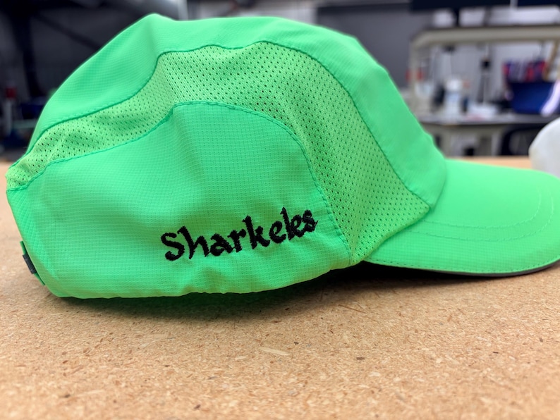 May include: A bright neon green baseball cap featuring a curved brim and breathable mesh side panels. The word Sharkeles is embroidered in black thread on the side of the cap. The hat is constructed from a lightweight, synthetic fabric material.