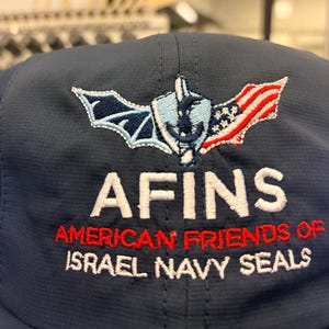 May include: A navy blue baseball cap featuring an embroidered logo with a bat wing, a shield with an anchor, and an American flag. Below the logo, the text reads AFINS, AMERICAN FRIENDS OF ISRAEL NAVY SEALS in white and red embroidered lettering.