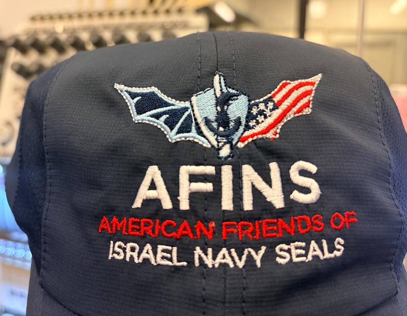 May include: A navy blue baseball cap featuring an embroidered logo with a bat wing, a shield with an anchor, and an American flag. Below the logo, the text reads AFINS, AMERICAN FRIENDS OF ISRAEL NAVY SEALS in white and red embroidered lettering.