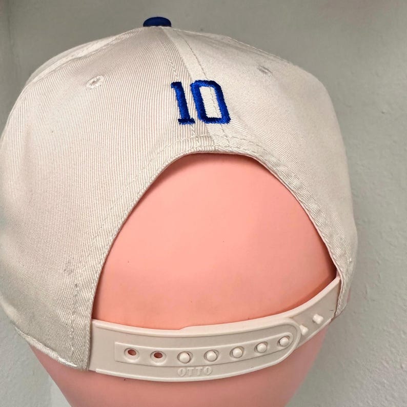 May include: A beige baseball cap featuring the number 10 embroidered in blue thread on the back panel. The cap includes a matching beige plastic snapback closure at the base with the word OTTO embossed on the adjustable strap mechanism.