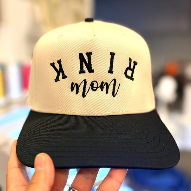 May include: A two-tone baseball cap with a cream-colored crown and a black flat brim. The front of the crown features black embroidered text that reads "KNINK" in a block font, with the word "mom" in a cursive script embroidered below it.