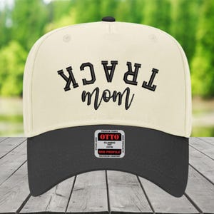 May include: A cream-colored baseball cap with a black brim featuring black embroidered text that reads TRACK MOM in a curved, block-letter style above a script font. A rectangular sticker on the brim displays the text OTTO PREMIUM FABRIC.